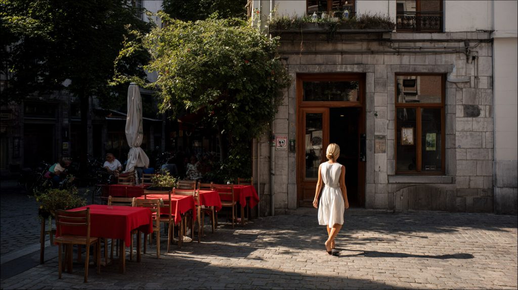 Vrouw in witte jurk loopt over een kinderkeitjesstraat naar de ingang van een gezellig restaurant met rood gedekte terrastafels